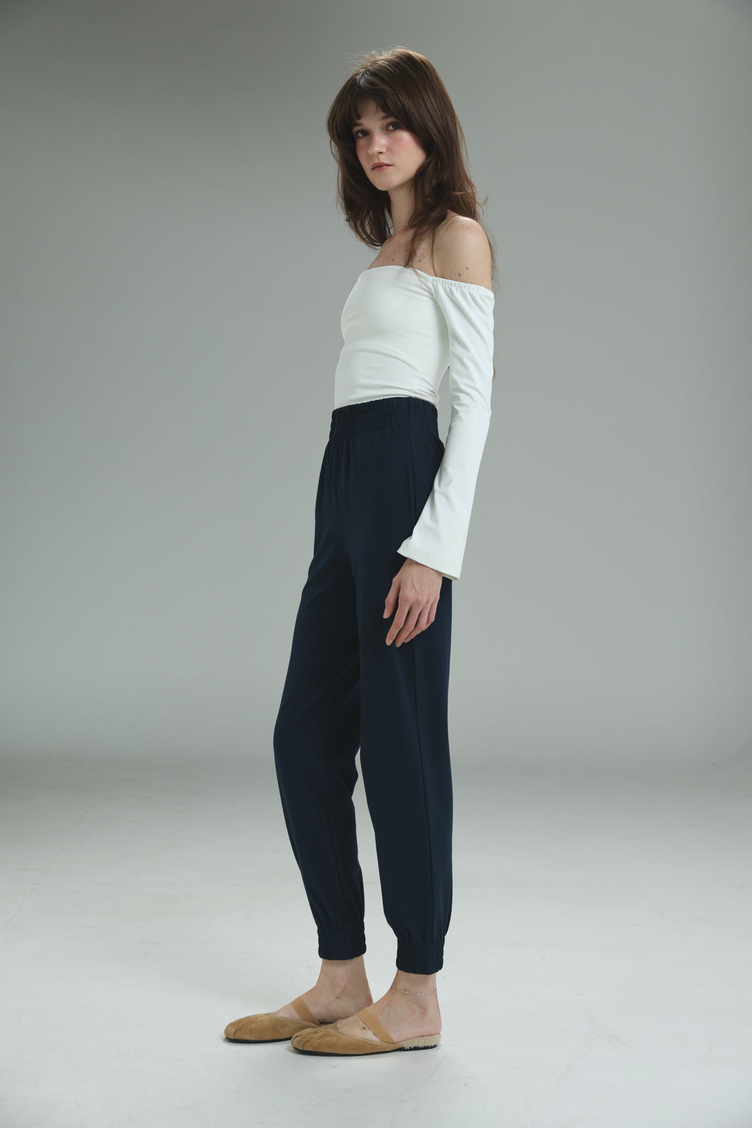 A white lycra off shoulder long-sleeved top worn with navy blue tapered jogger pants and tan flat mules, front view