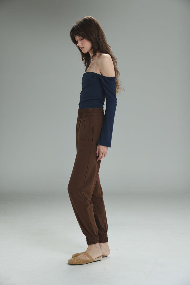 A woman wearing a lycra navy off shoulder long-sleeved top shown from the front with brown tapered pants and tan sandals