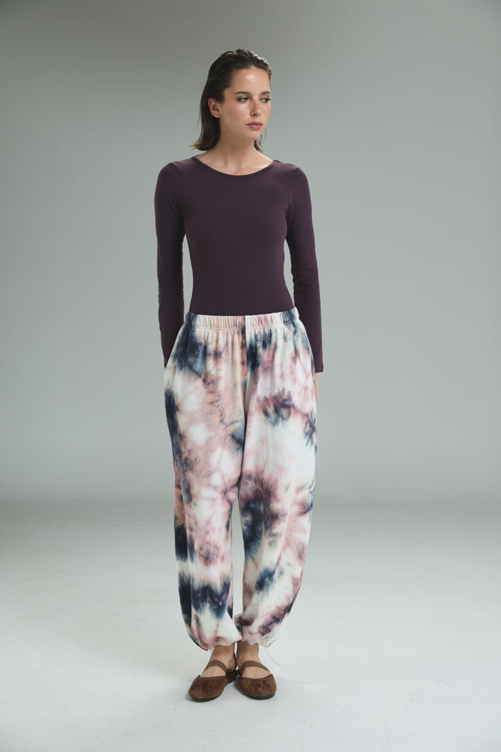 The model wears a lycra purple open back bodysuit paired with flowing pastel pink and purple tie-dye harem pants