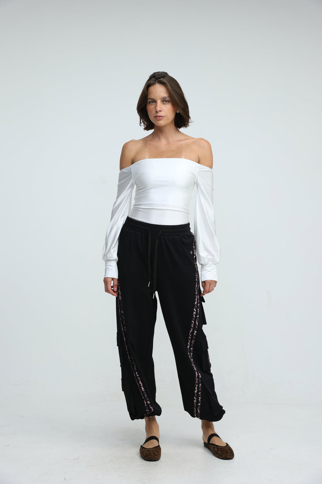 A woman wearing port black joggers with decorative side stripes and a white off-the-shoulder long sleeve top