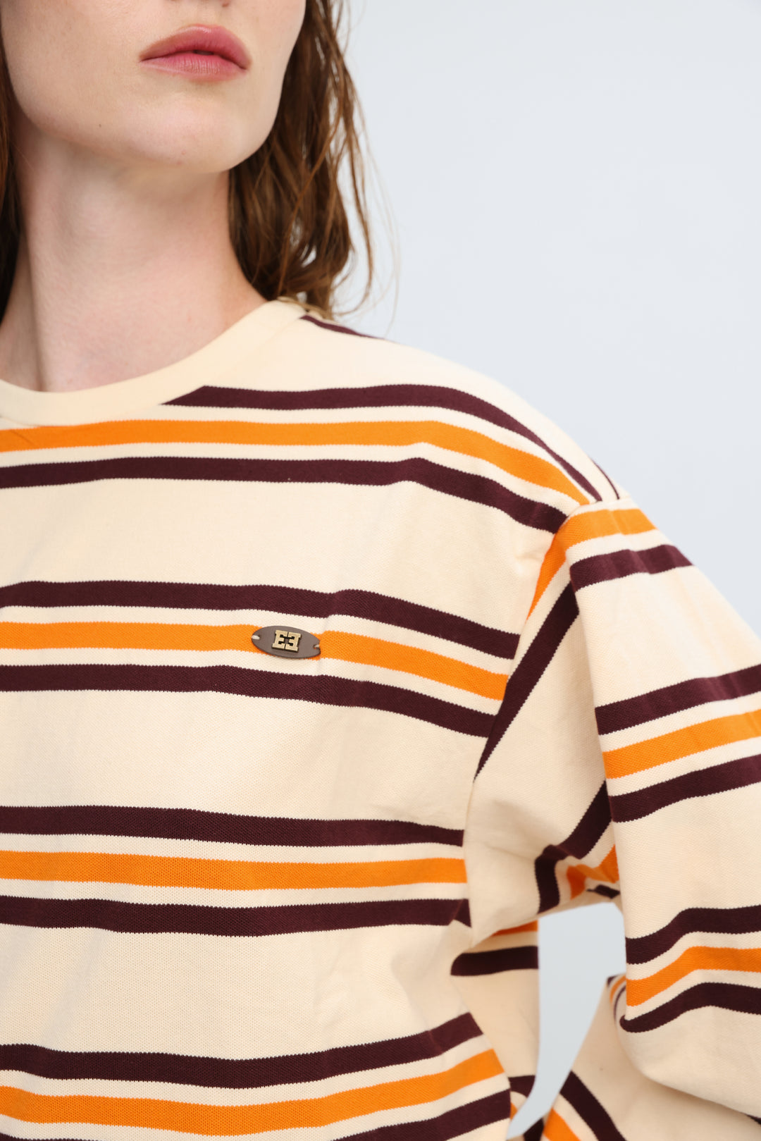 Cream-colored sweatshirt with bold orange and dark brown horizontal stripes and small metallic logo patch on front