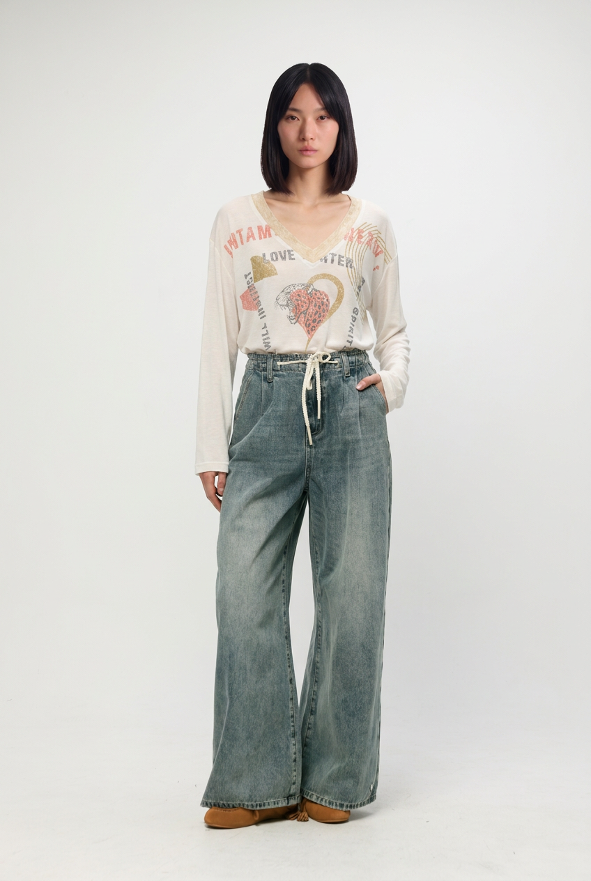 A woman wears a cream long-sleeved graphic tee with miami and heart designs paired with star 461 blue denim wide-leg pants