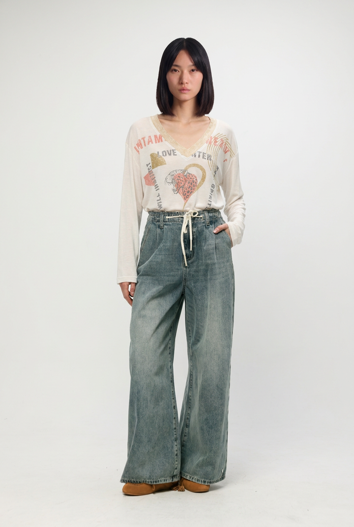 A woman wears a cream long-sleeved graphic tee with miami and heart designs paired with star 461 blue denim wide-leg pants