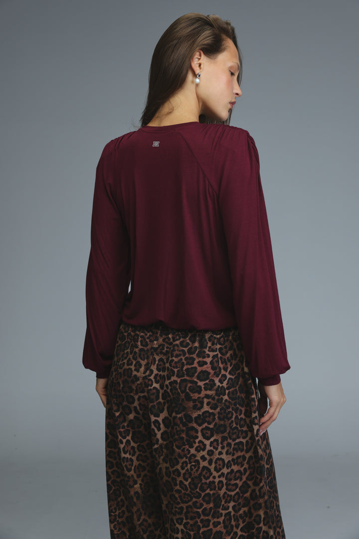 A woman wearing a maroon long-sleeved wine padded shirt with puffed sleeves and subtle back detail visible from the side