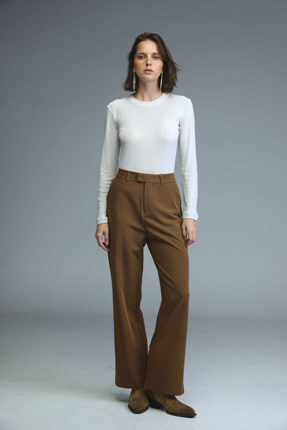 A woman wearing mid rise tailored brown wide-leg trousers paired with a white long-sleeved top and brown suede shoes