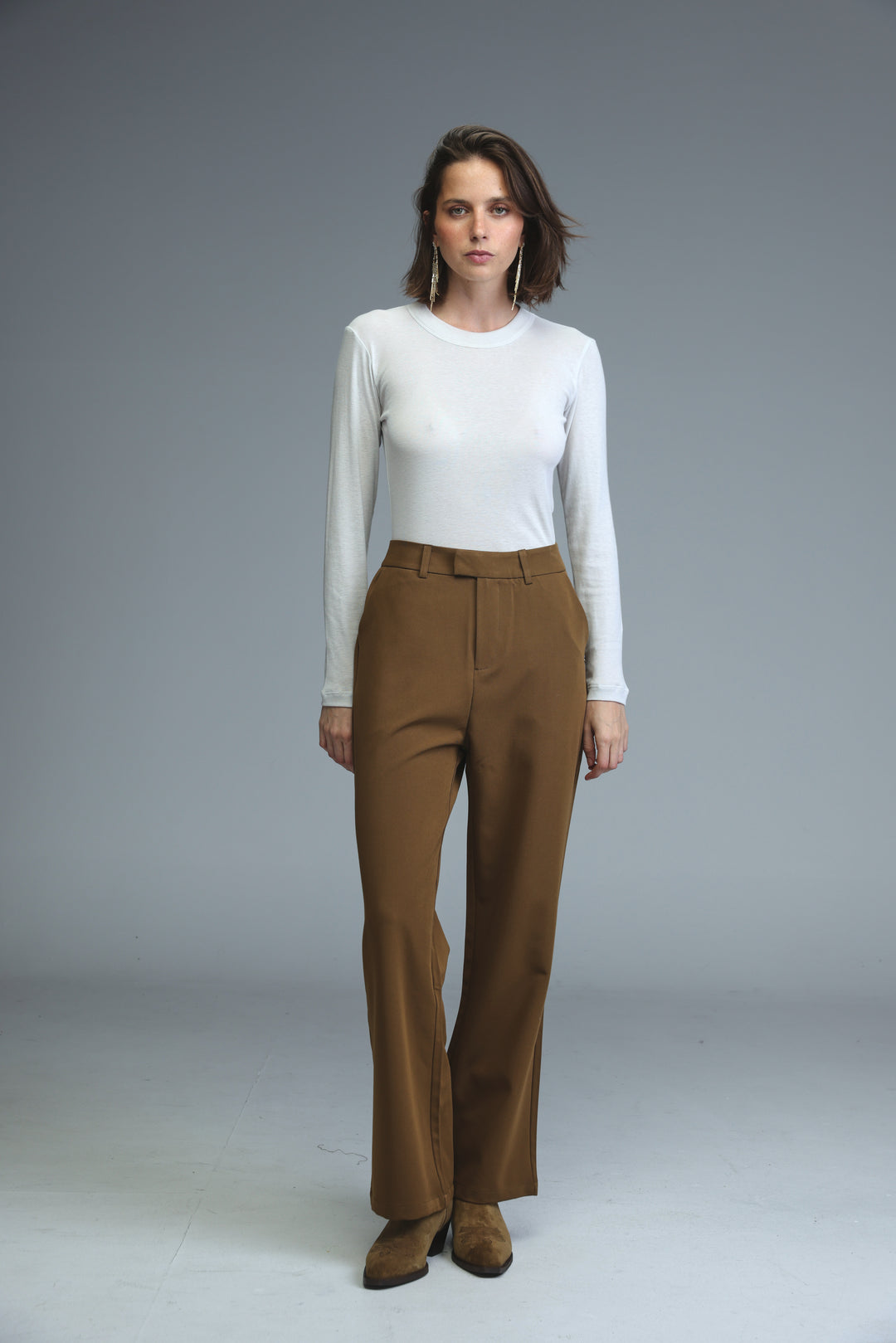 A woman wears mid rise tailored brown trousers paired with a white long-sleeved top, front view