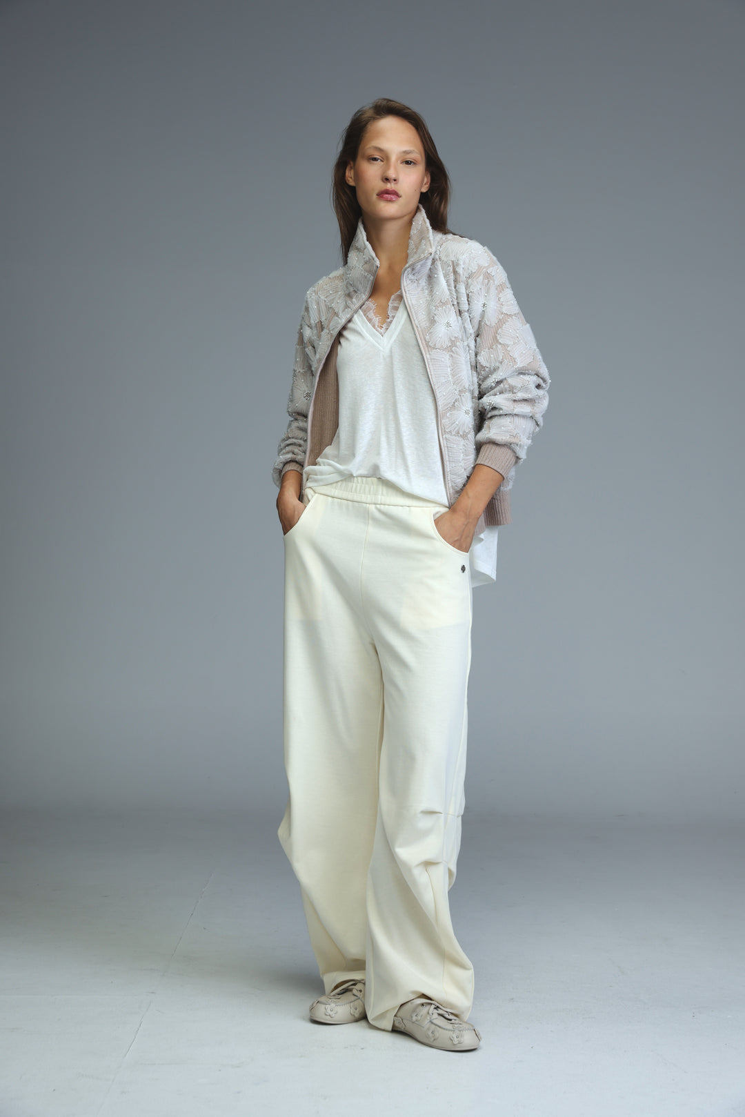 A woman wearing a light gray Blossom Stone quilted bomber jacket with a subtle snakeskin pattern and white trousers