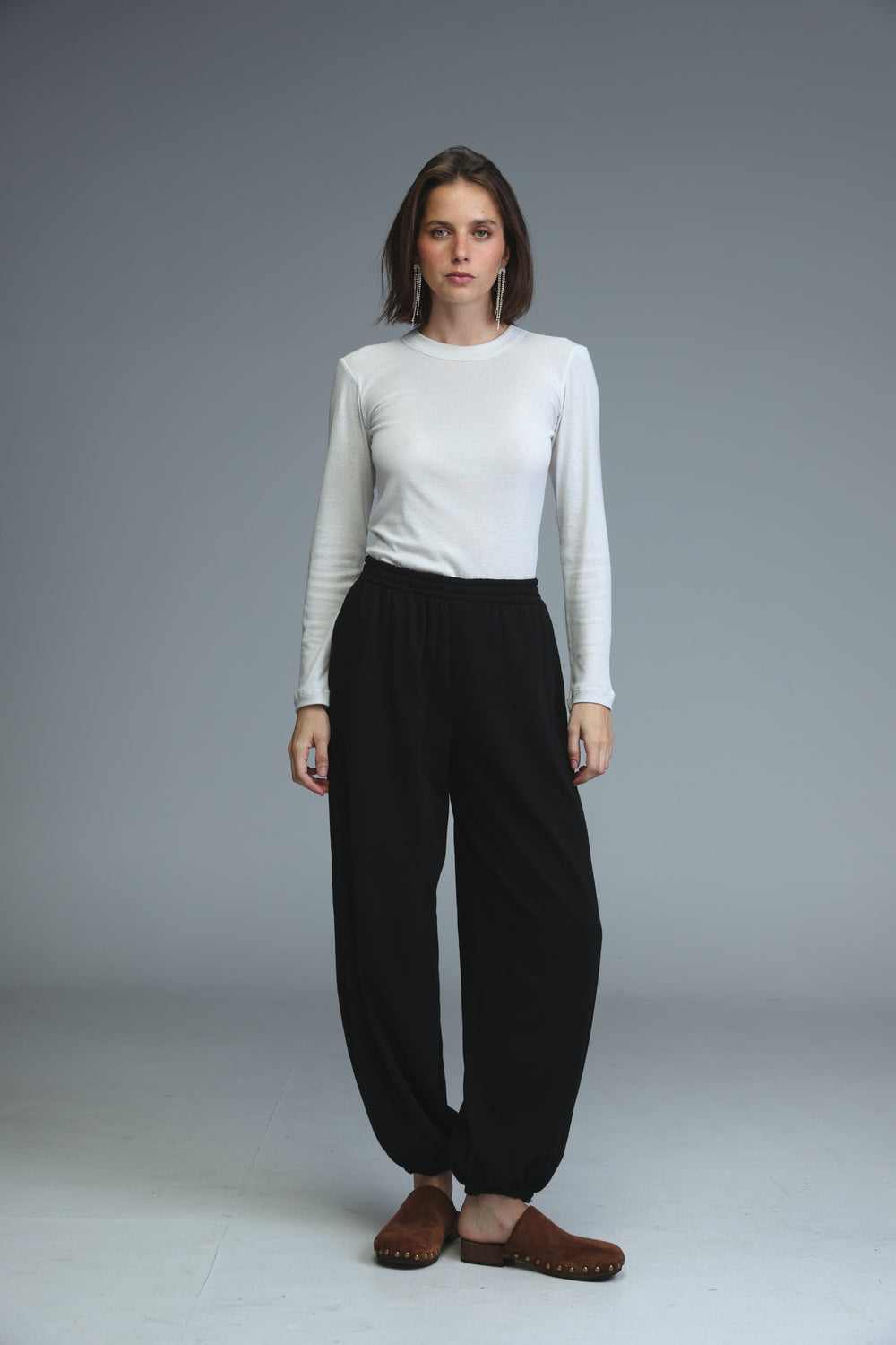 A woman wears black joggers with a white long-sleeved top and brown suede mules in a front view