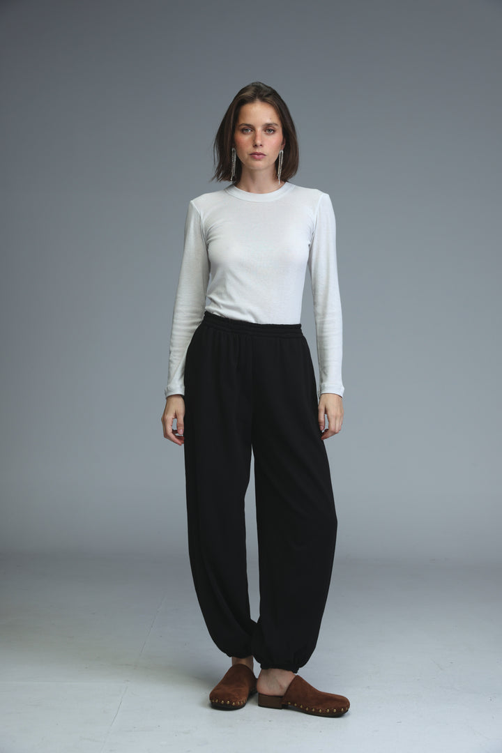 A woman wears black joggers with a white long-sleeved top and brown suede mules in a front view