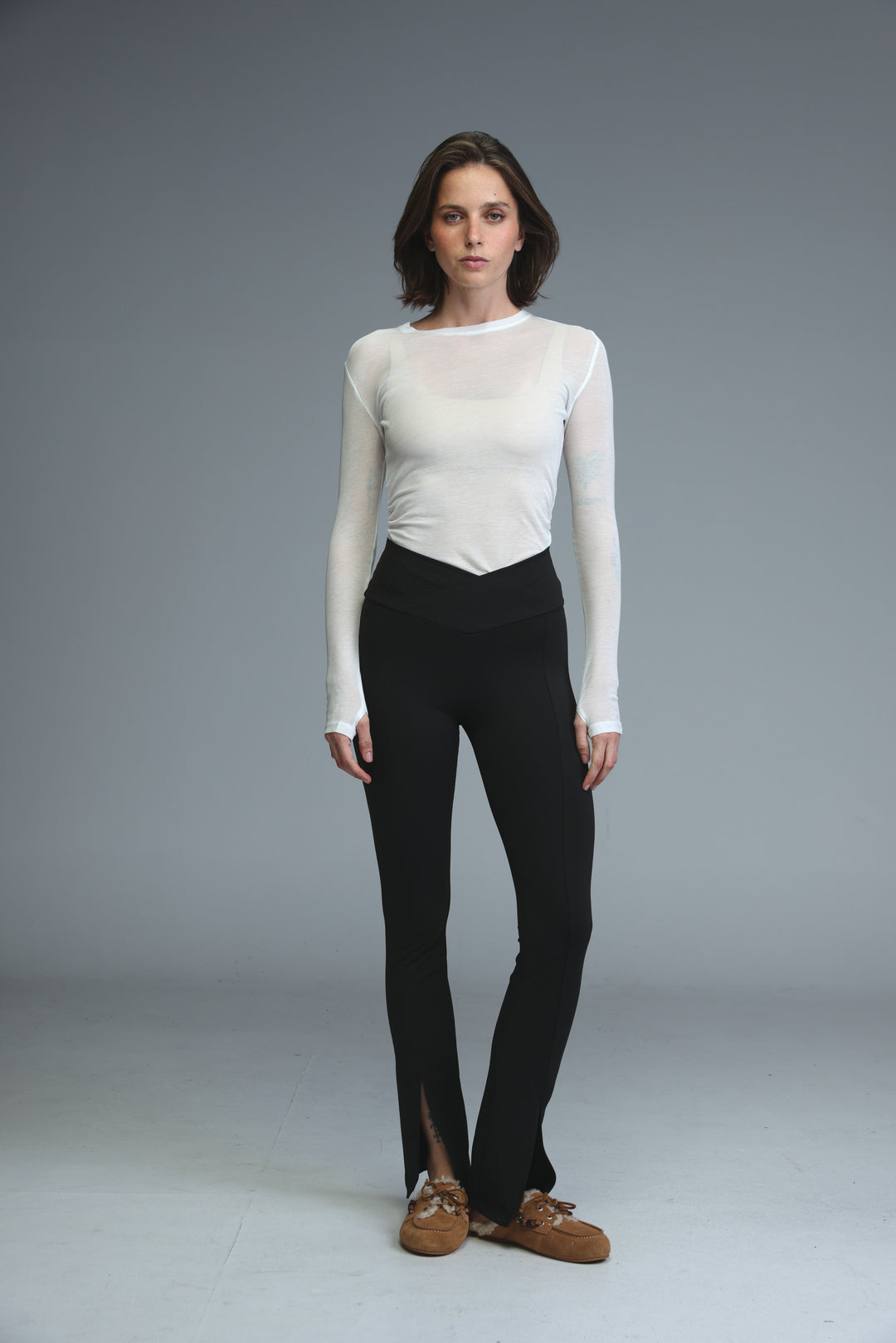 A woman wearing front slit black leggings paired with a white sheer top and brown suede moccasins in a side view