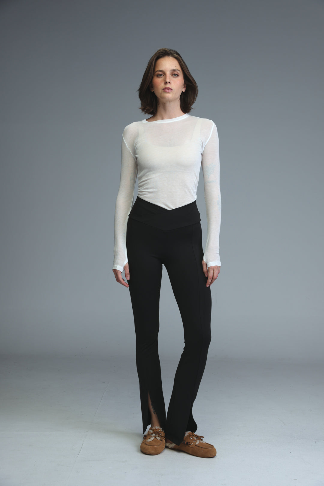 A front view of black front slit leggings paired with a white sheer long-sleeved top and brown suede loafers